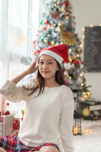 Portrait photo of a young beautiful friendly asian female lady with Christmas santa hat posing in front a nice decorated christmas tree as background in her room