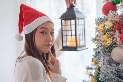 Portrait photo of a young beautiful friendly asian female lady with Christmas santa hat holding an outdoor lantern candlelight in front a nice decorated christmas tree