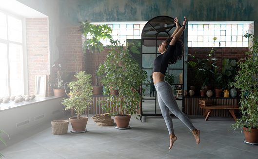 Slim Afro-American woman stretching at home or fitness studio in the morning. Studio decorated with plenty house plants, big windows and brick wall. Concept of happy and conscious healthy lifestyle.