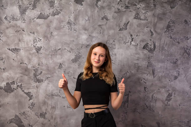 portrait teenager girl looking at camera thumbs up hands at grey textured background. confident teen girl 12-13 years old in stylish black wear, posing, showing good gesture. copy text space - 12 13 years pre adolescent child female blond hair imagens e fotografias de stock