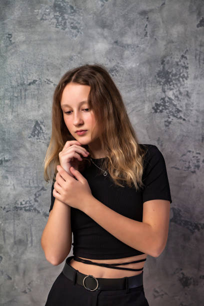 portrait sad teen girl in black clothes thinking looking down at grey textured background. pensive thoughtful teenager girl 12 years old posing in studio. fashion style concept. copy text space - 12 13 years pre adolescent child female blond hair imagens e fotografias de stock