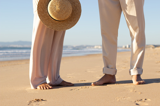 Closeup of couples legs standing barefoot on wet sand at seashore. Woman with straw hat in hand and man in beige trousers enjoying vacation. Sea background. Vacation, leisure concept