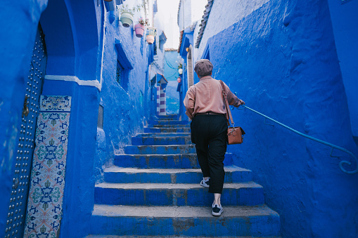 Rear view Asian Chinese female tourist walking into alley in Chefchaouen admiring the surrounding blue wall
