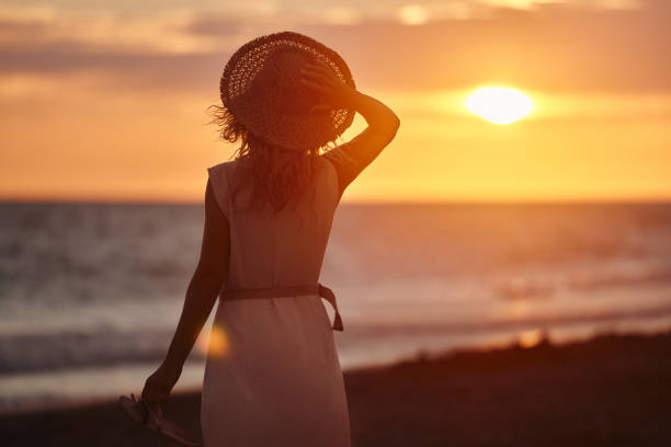 Rear view of a woman with sun hat standing on the beach and looking at sunset above the sea. Copy space.