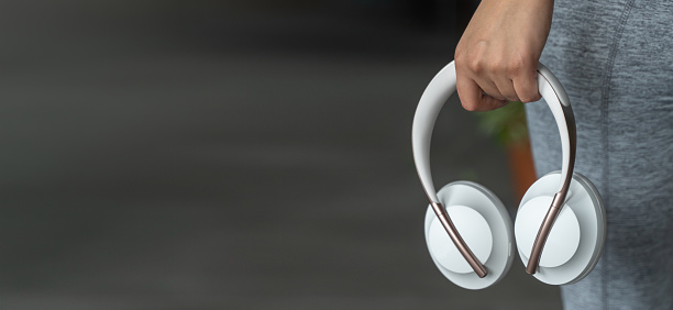 A close-up shot of a young female professional listening to music through wireless headphones, with eyes closed in an urban setting. She is wearing smart-casual winter clothing and her hands reach to the headphones.