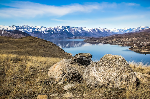 A spectacular view of Lake Chelan, Washington, looking westward from Chelan Butte.  Along the right side of the lake the nearby town of Manson can be seen with it's buildings dotting the right side of the shoreline.  Snow capped mountains majestically rise up in the distance while a strong, rock formation surrounded by winter grass dominates the foreground.