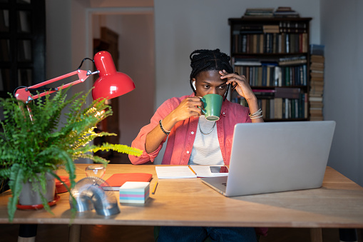 Tired African male student studying remotely from home until late, drinking coffee, sleepy black guy trying to concentrate while watching online course on laptop computer in evening. Learning fatigue