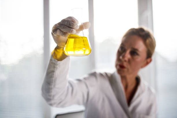 Selective focus shot of serious mid adult female laboratory technician, wearing protective gloves, holding erlenmeyer flask with yellow liquid, swirling it around and analyzing it. Focus on foreground.