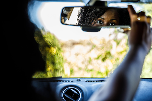 Over the shoulder view of young Black woman sitting in the driver's seat of her new car and adjusting the rear view mirror.