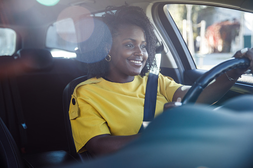 Portrait of charming young Black woman sitting in driver's seat in her car and smiling and enjoying the ride to her location. Seen through windshield.