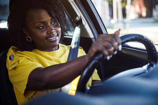 Portrait of charming young Black woman sitting in driver's seat in her car and smiling and enjoying the ride to her location. Seen through windshield.