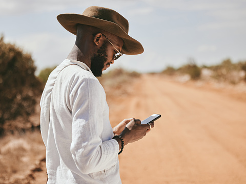 Dessert dirt road, black man and phone text in nature on a vacation lost and waiting for a lift. 5g internet, mobile and web app usage of a person from Madrid wait for a taxi to travel in summer