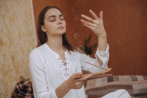 Peaceful woman with burning incense stick smelling smoke during aromatherapy while relaxing at home