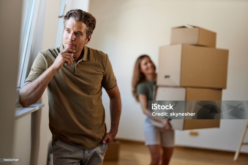 I know I'm supposed to do something, but I don't know what! Thoughtful man thinking what he was supposed to do while his wife is struggling with boxes in the background. Copy space. Ignorance Stock Photo