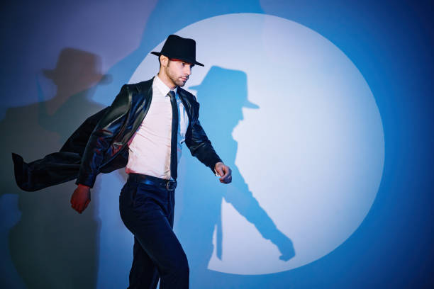 Handsome man in black coat and hat walking like superhero in the spotlight with copy space. Noir film style. Private detective, spy, investigation concept