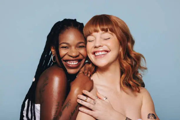 Cheerful young women with different skin tones smiling while standing together. Two happy young women feeling comfortable in their own skin. Body confident young women standing topless in a studio.