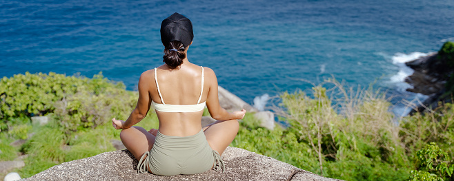 Back of young Asian woman meditating on the cliff of mountain with a sea view. Beautiful female working out outdoors