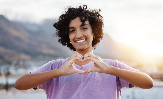 Hand, heart and woman showing love sign in a city, travel, freedom and happy student. Portrait, smile and finger gesture with cheerful indian tourist enjoying foreign exchange programme in Mexico