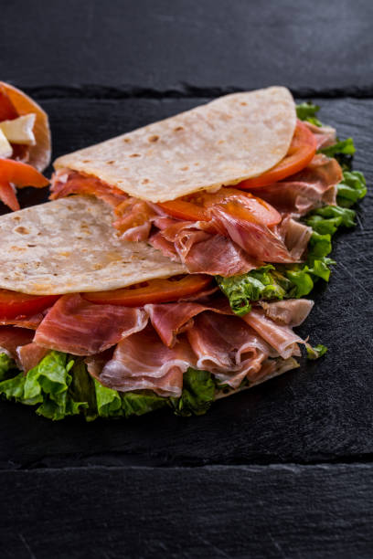 istock Piadina with prosciutto, tomatoes, lettuce, cut in half on a black, stone background. 1445134048