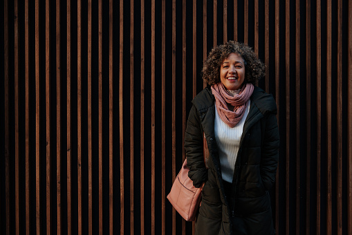 A mature African American woman is standing outside in front of a wall, looking at the camera and smiling in a winter jacket and a scarf with copy-space.