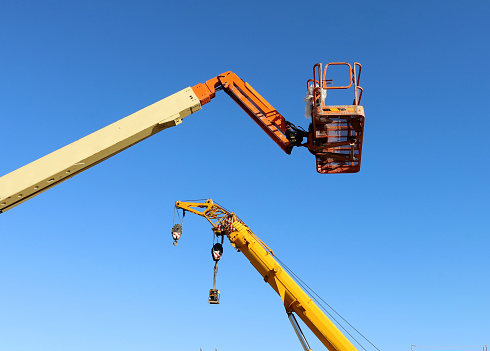 Bucket of cherry picker with a boom of mobile crane on behind, against blue sky. Background for copy space