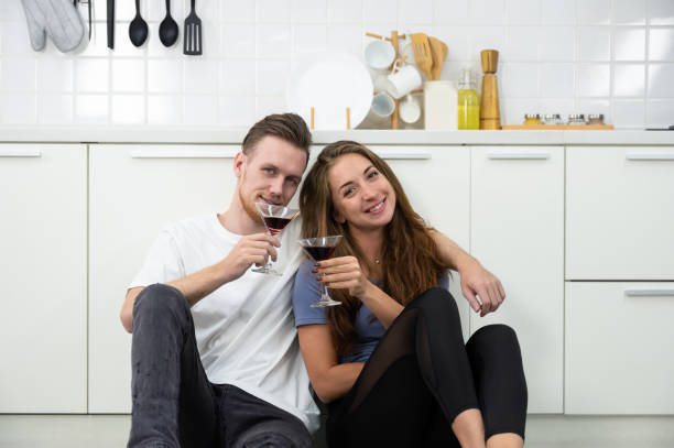 jeune amoureux du couple assis sur le sol de la cuisine et tenant des verres de cocktails ou de vin, s’amusant et appréciant de boire à la maison. - placard holding celebration women photos et images de collection