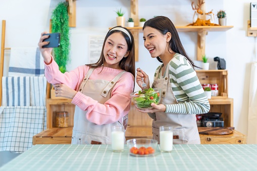Photo of two young beautiful asian woman lady happy selfie video call and enjoy drinking milk and eating salad with cherry tomato in kitchen of their house for diet and lose weight for healthy lifestyle