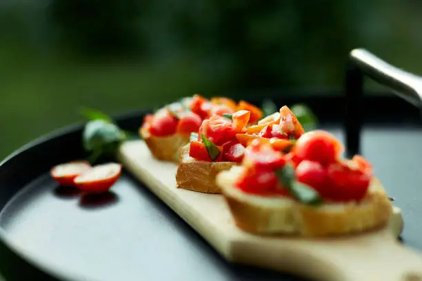 Italian bruschetta with fresh red tomatoes and basil herbs on a wooden board outdoor on the yard, backyard at home, sunlight. Classic Italy food, antipasto