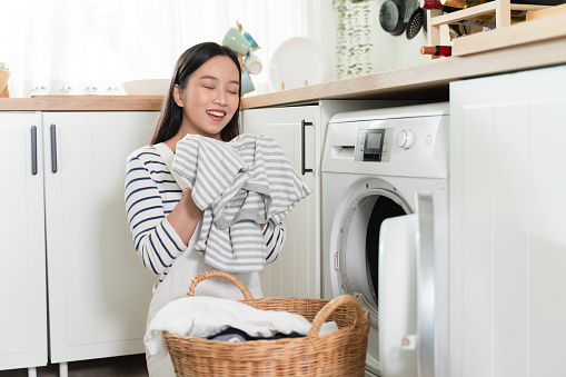 Young happy Asian woman smelling and doing laundry at home. Beautiful smiling girl putting clothes in front loading washing machine. Housework and chores concept