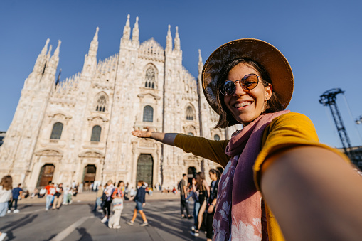 Beautiful young female tourist having a video call using smart phone in front of the cathedral in Milan, Italy.