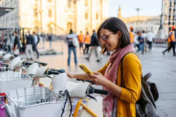 Beautiful young female tourist renting an electric bike  In Milan, Italy using a smart phone app.