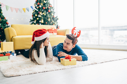 Young happy Asian woman wearing a Santa Claus hat with her boyfriend with a Christmas gift while lying down on the carpet with a Christmas tree in the background. Photo with copy space.