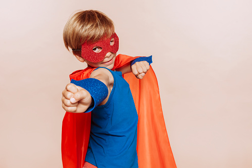 Funny little superhero boy wears red cape and mask, with raised hand at studio isolated over beige background. Childhood, fantasy, children playing concept.