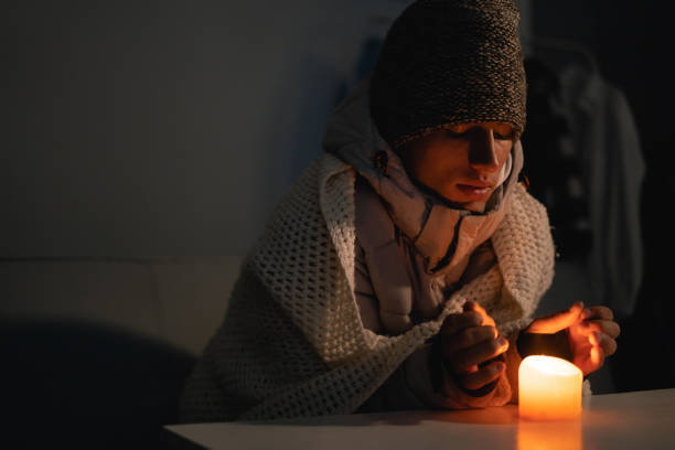 istock A young man dressed in a warm winter hat at home at a table and warms his hands from a burning candle. Blackout city, electricity off, energy crisis or power outage, concept image. 1445175303