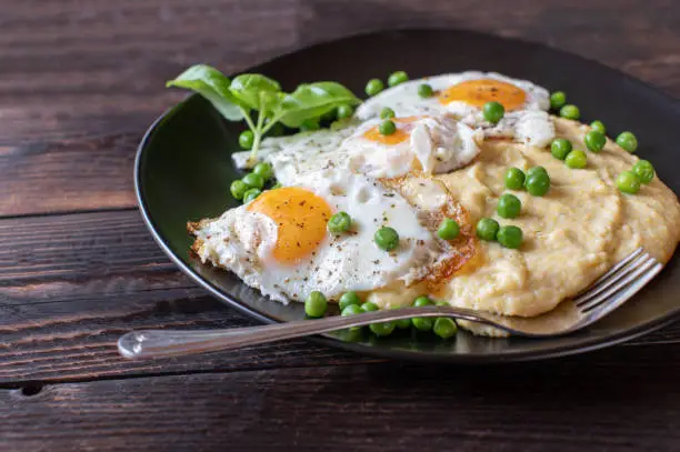 Delicious vegetarian meal for lunch or dinner with fried eggs, sunny side up, creamy polenta and green peas on a brown plate on brown wooden background. Closeup and front view