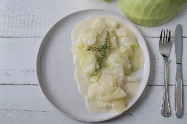 Fresh and homemade cooked white cabbage with a delicious bechamel sauce  isolated on a plate for dinner or lunch side dish. Served on white wooden background from above