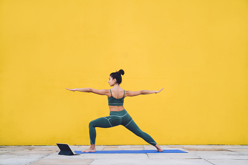 Side view of focused calm female in sportive outfit concentrating in warrior pose on yoga mat with yellow background behind