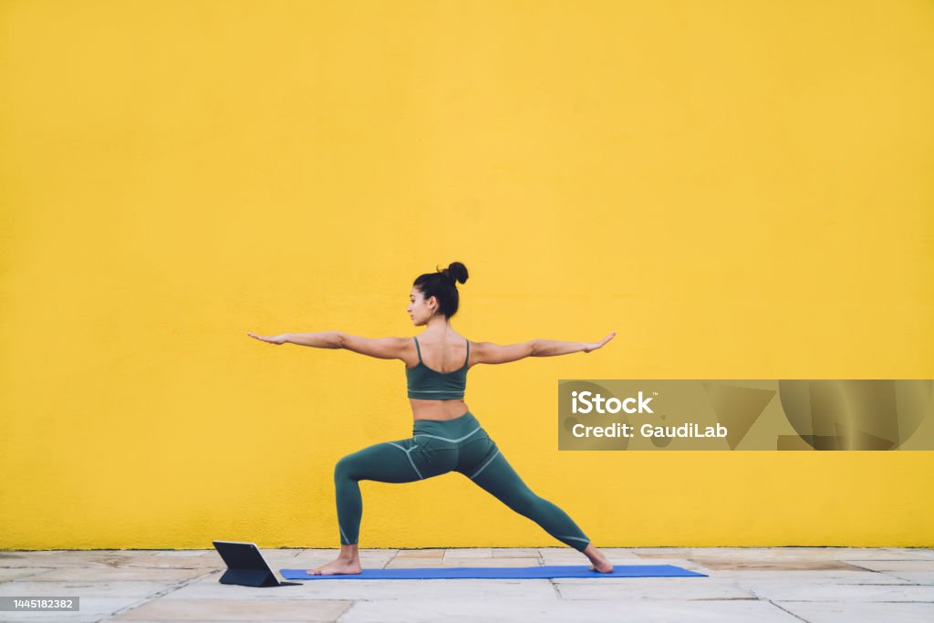 Fit woman practicing yoga with tablet outside Side view of focused calm female in sportive outfit concentrating in warrior pose on yoga mat with yellow background behind Yoga Stock Photo