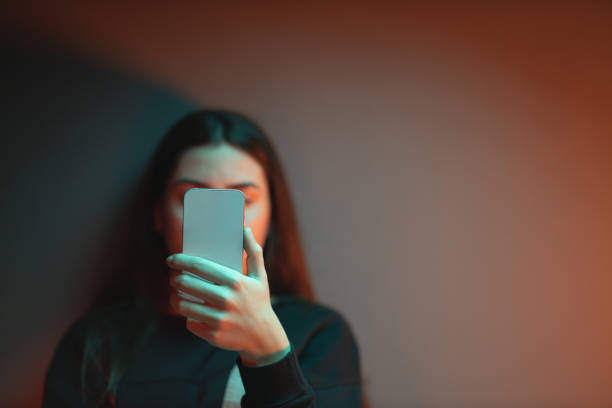 The social media overuse Close-up of a depressed teenage girl sitting on the bed in her room lightened with orange and teal neon lights and scrolling social media. harassment stock pictures, royalty-free photos & images