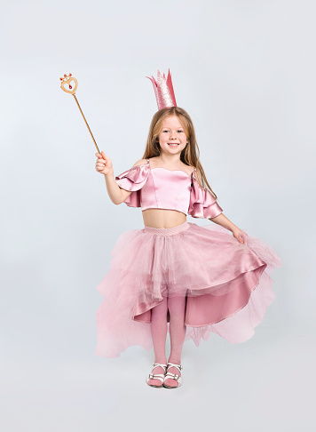 Cute girl in fairy dress with pink crown and magic wand on light grey background. Little princess