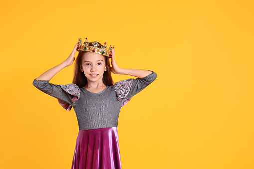Cute girl in dress and golden crown with gems on yellow background, space for text. Little princess