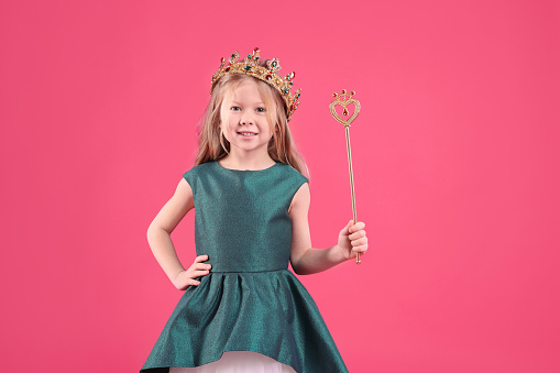 Cute girl in fairy dress and golden crown holding magic wand on pink background. Little princess