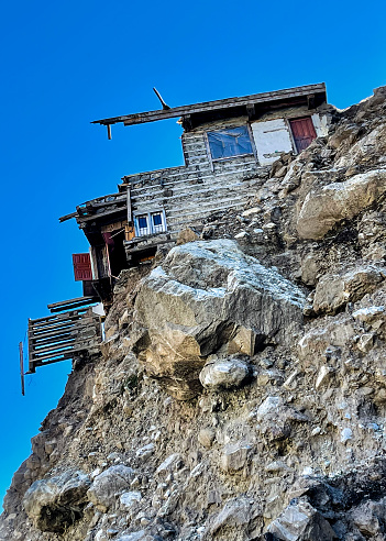 A damaged house hanging by the cliff in Pashmal town in Swat Valley, Pakistan on 26 October 2022, a few months after Pakistan’s worst flooding in a decade caused by climate change. Swat is the country’s most popular tourist destination and is often called the Switzerland of Asia.  It was also the center of an ancient civilization of Ghandara Kingdom, and a house of numerous Buddhist sites, which makes Swat one of the destinations for Buddhist pilgrimage. 

The photo shows a damaged house which some parts were washed away by the flood and only the other half of the house left hanging by the eroded mountain. The photo was taken during the sunny day with a blue sky in the background. The mountain was covered by green grass before the flood and now left with brownish sand and stones. The roof the house is still there, and there is a window and door in the image.