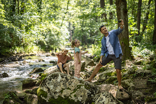 Father with two kids, son and daughter standing on rock and taking selfie with 
wearable camera by the forest stream