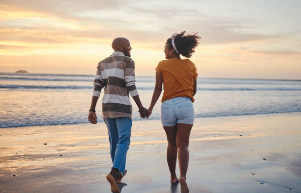 istock Black couple, holding hands and on beach with sunset for romance, celebrate relationship or holiday together. Love, happy man and woman bare feet, relax or smile on romantic seaside vacation or break 1445191976