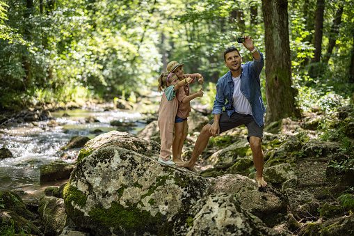 Father with two kids, son and daughter standing on rock and taking selfie with 
wearable camera by the forest stream
