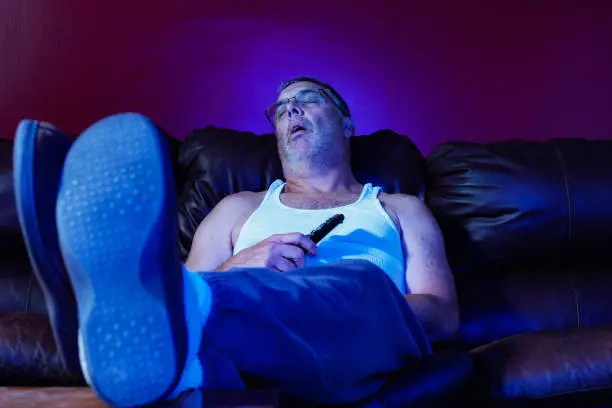 A middle aged Caucasian man with TV remote on leather sofa asleep while binge watching TV at night.