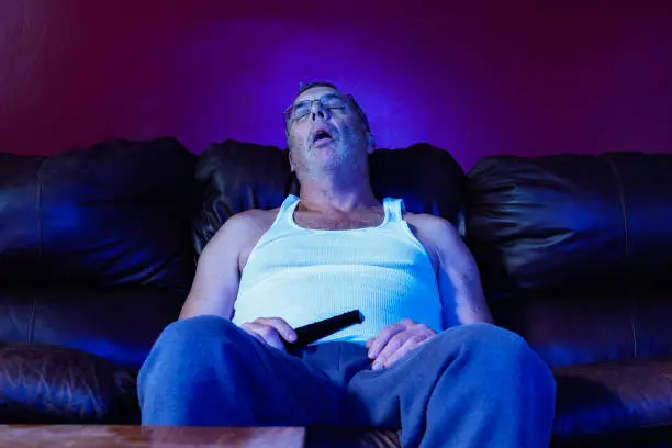 A middle aged Caucasian man with TV remote on leather sofa asleep while binge watching TV at night.