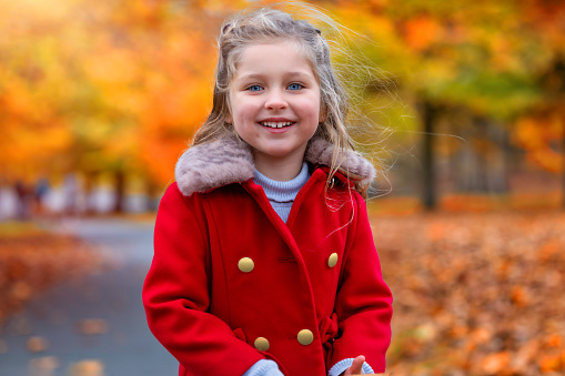 Portrait of a cute, blonde girl in a red winter coat standing in a park with colorful leaves during autumn time