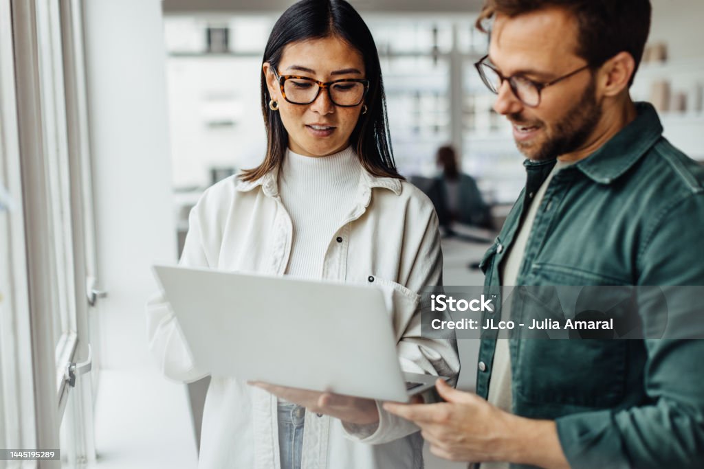 Business professionals standing in an office and using a laptop Business professionals standing in an office and using a laptop together. Two young business people having a discussion in a workplace. Office Stock Photo